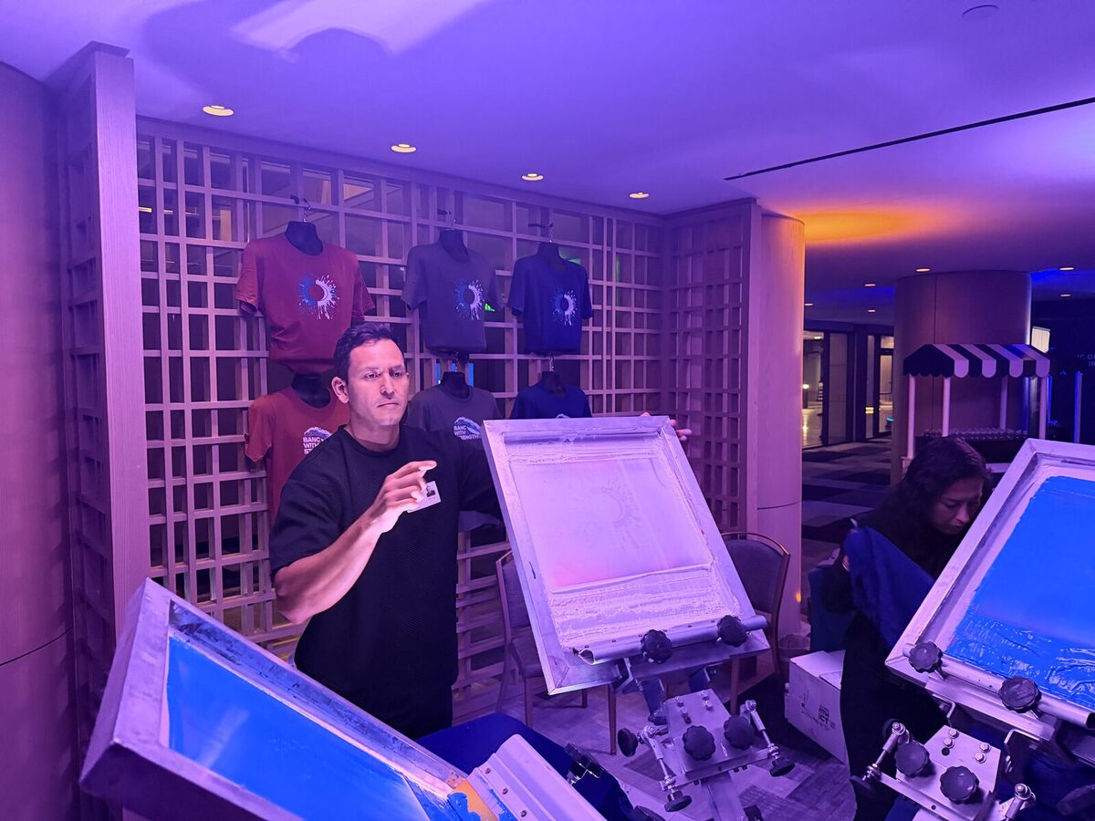 Printer holding a screen at a live hat-and-shirt station with finished tees displayed on a lattice wall