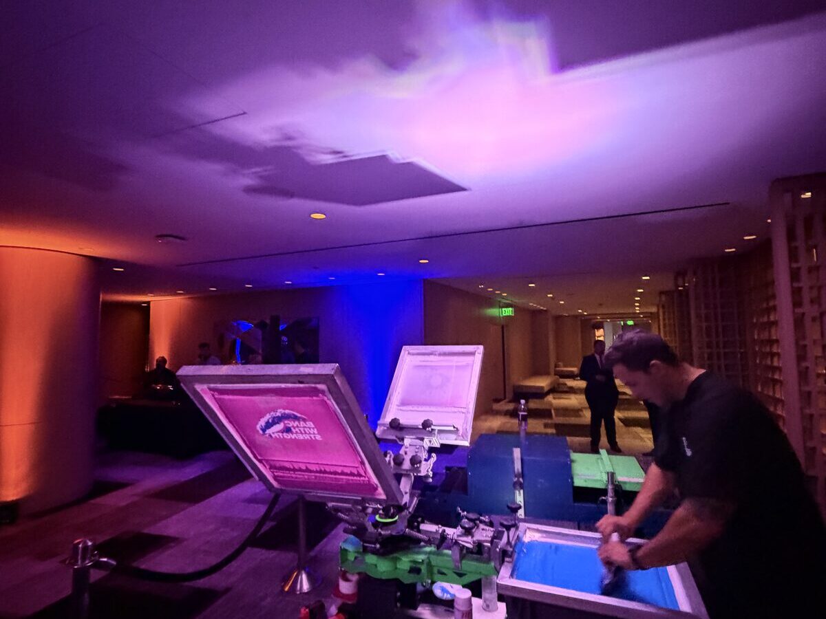 A printer inking a screen on press, lit by warm and blue event lighting at an evening activation